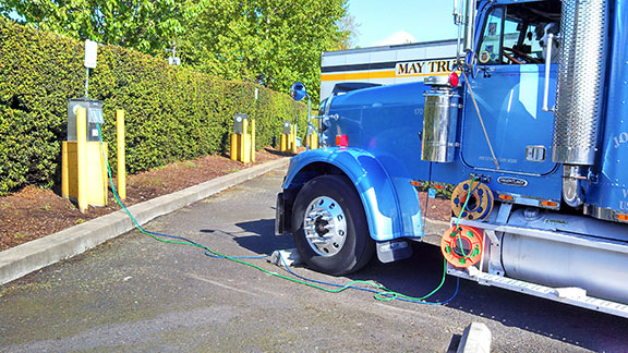 This figure is a photo of a sleeper cab tractor parked at a modern truck stop. The tractor is plugged into a nearby power outlet which allows the cab to be heated or air conditioned while the driver is resting inside.